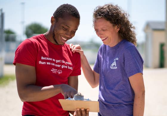 A person in a red shirt with the words "Because we get far more than we can ever give back" holding and pointing at a clipboard while a person in a purple shirt smiles down at the clipboard. 